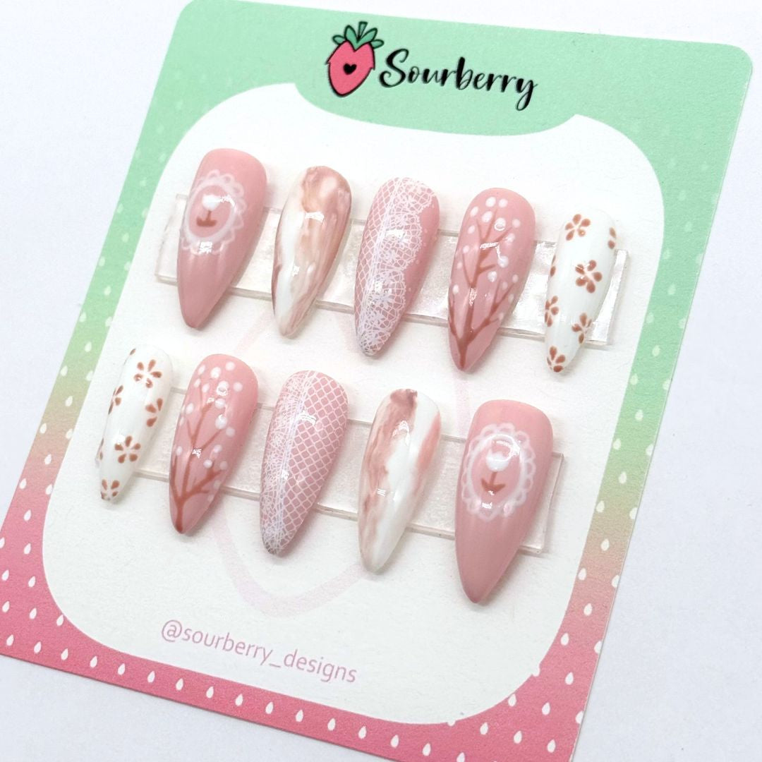An image of a Sourberry brand Countryside Sonnet nail set on a pink and green mounting card. The nail sets are beige and white with lace, marble, and floral details.