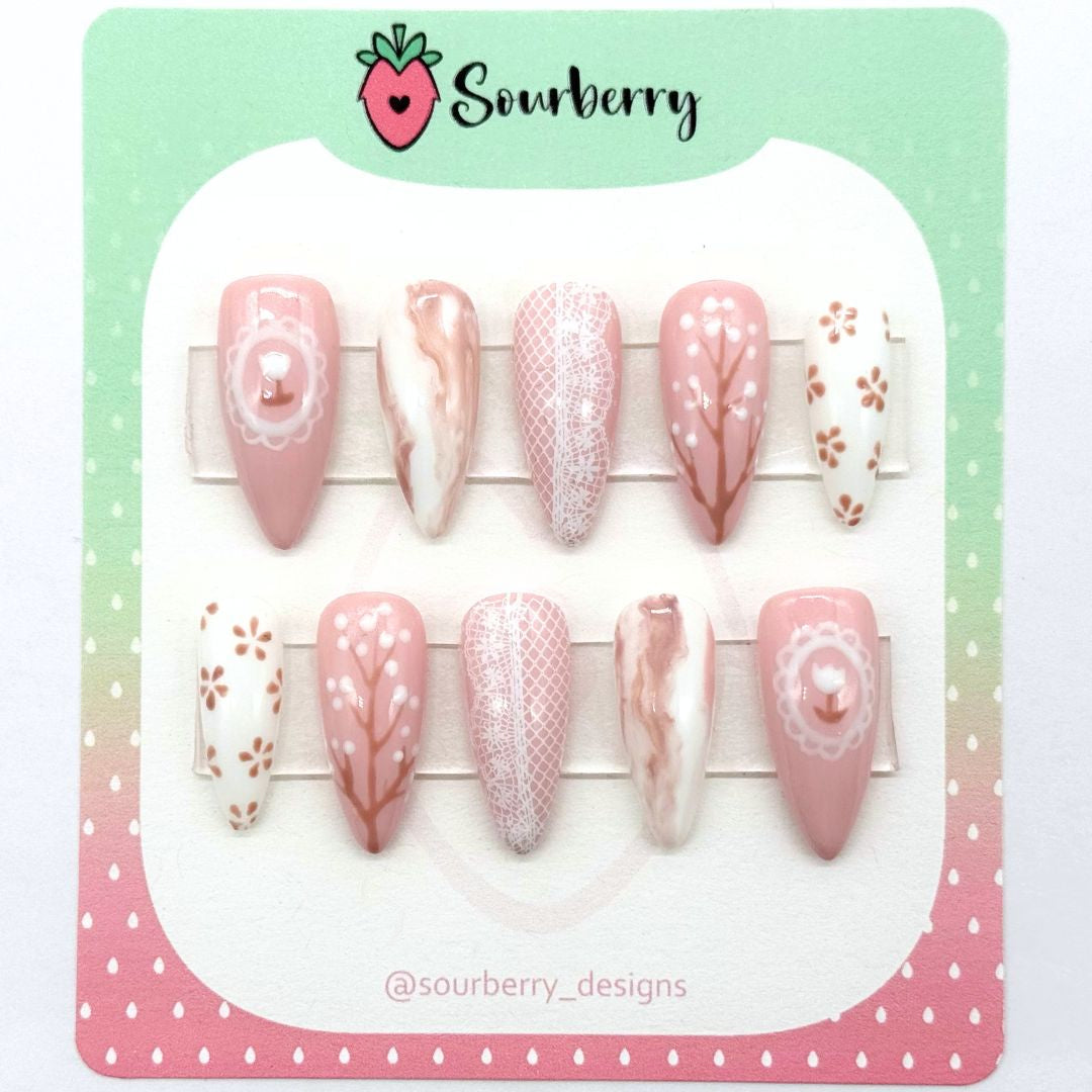 An image of a Sourberry brand Countryside Sonnet nail set on a pink and green mounting card. The nail sets are beige and white with lace, marble, and floral details.