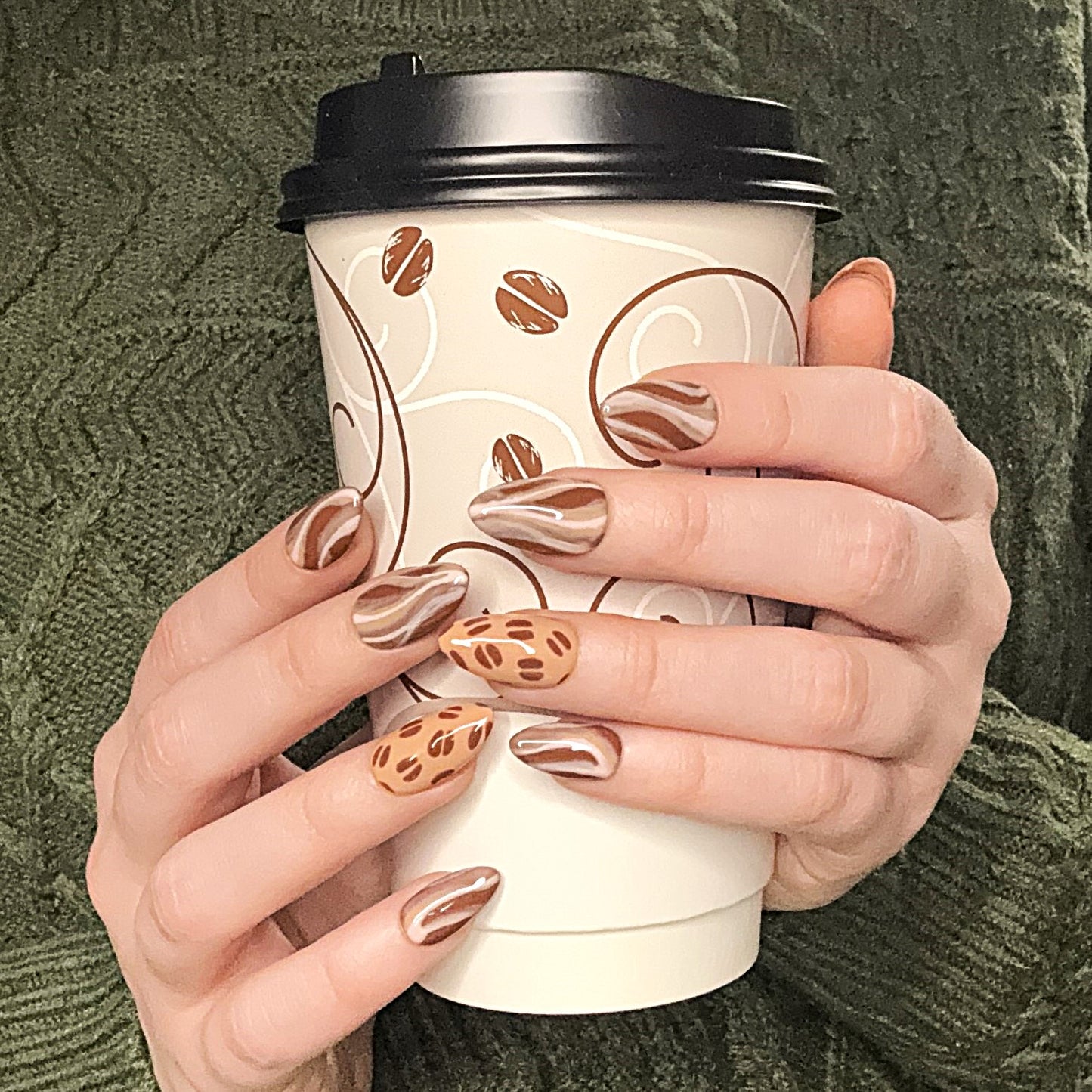 Cafe Latte nails on two hands holding a coffe cup. The background is a green textured sweater.