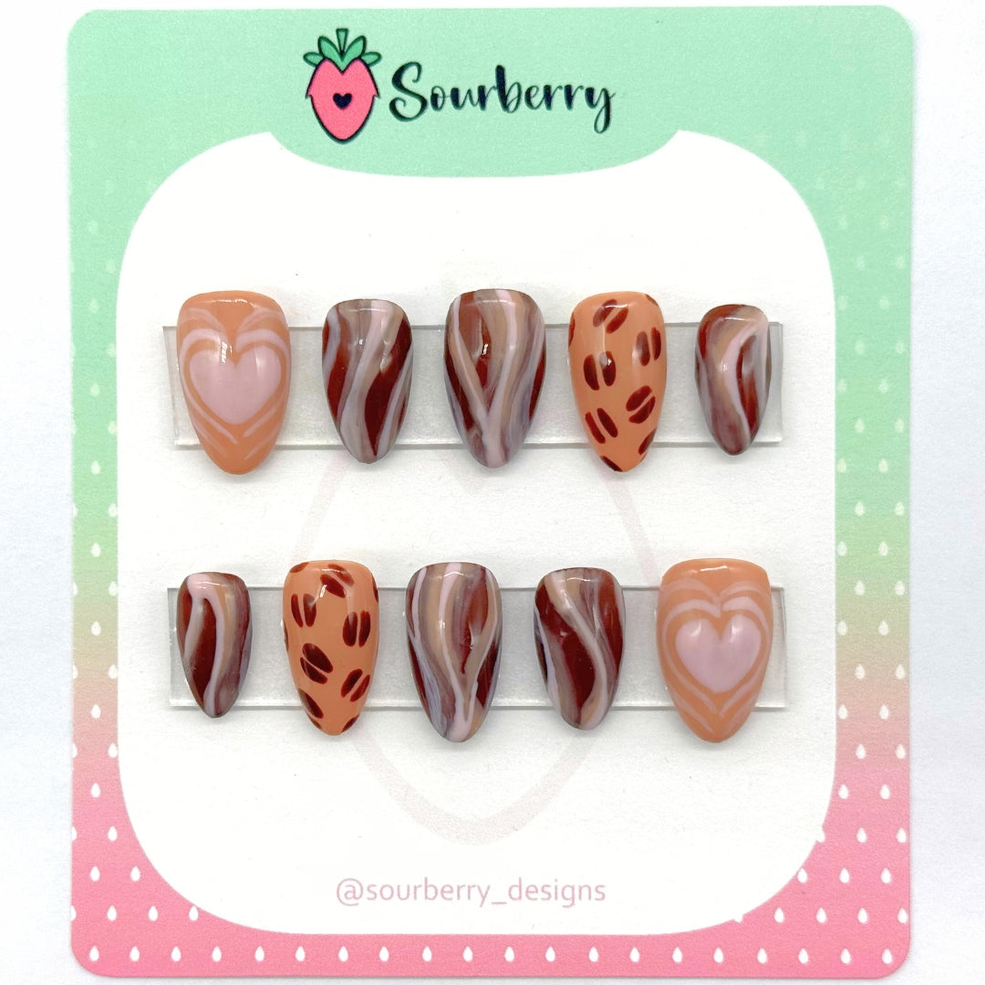 A picture of Sourberry brand "Cafe Latte" press-on nails mounted on a green and pink card. The thumb nails have a latte heart pattern on them. The ring finger nails have a coffee bean pattern. The other nails have a brown and white swirl.
