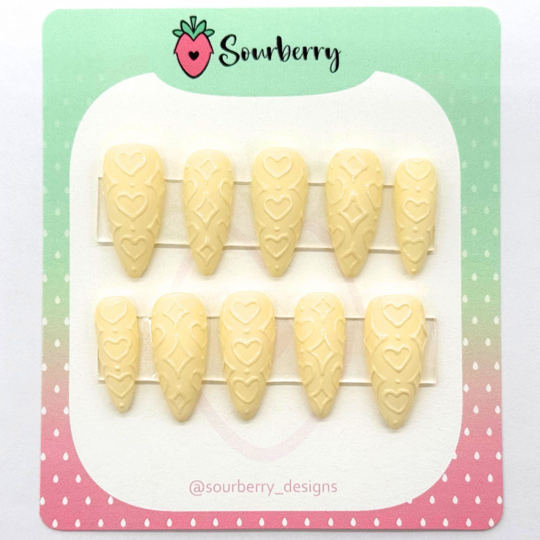 A picture of Sourberry brand "Powder Puff" press-on nails mounted on a green and pink card. The nails are light yellow with 3D heart and star patterns.