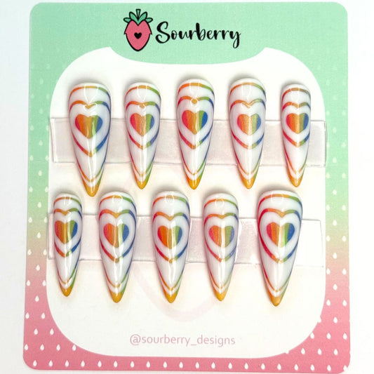 Rainbow Love Bomb nail set. This set has a white background with glittery rainbow heart patters on them. 