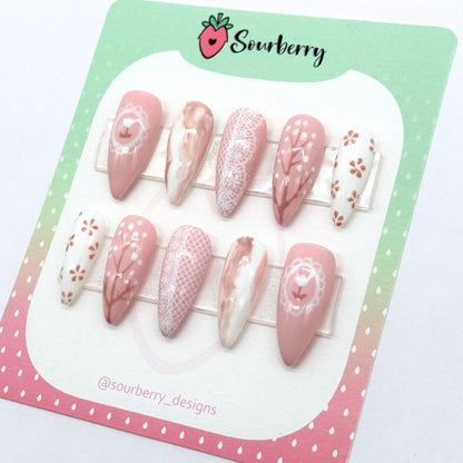 An image of a Sourberry brand Countryside Sonnet nail set on a pink and green mounting card. The nail sets are beige and white with lace, marble, and floral details.