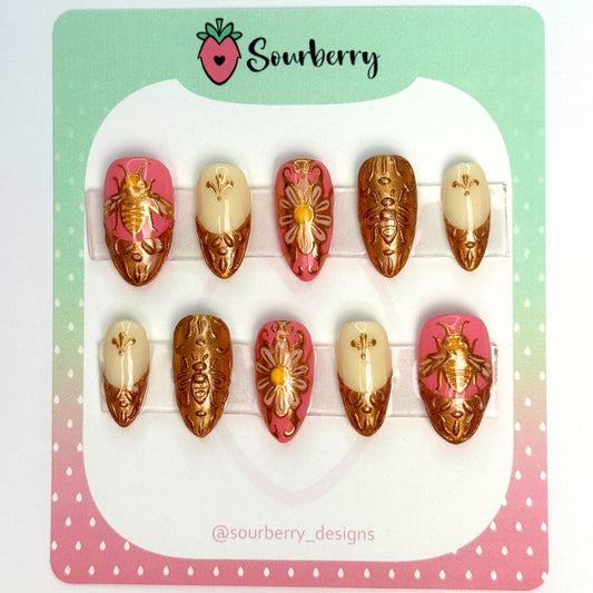 Royal Honey nail set. The thumb nails have a bee on a pink background with a golden french tip. The Pinky nail and index nail have a golden french tip and tiny gold details. The middle nail has a white flower covered in gold detailing. And the ring nails are completely gold with 3D golden bumblebee designs.
