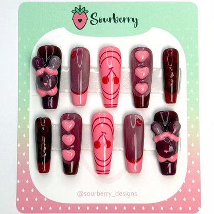 Cherry Cola nails. The thumb nail has a 3D sparkly red rabbit on it. The index nail is red and shimmery with a red kiss mark and a pink french tip. The middle nail is pink with a red cherry design. The ring nail is shimmery red with a dark red french tip and 3D pink hearts. The pinky nail is dark red with "cola" written on it and a red french tip.