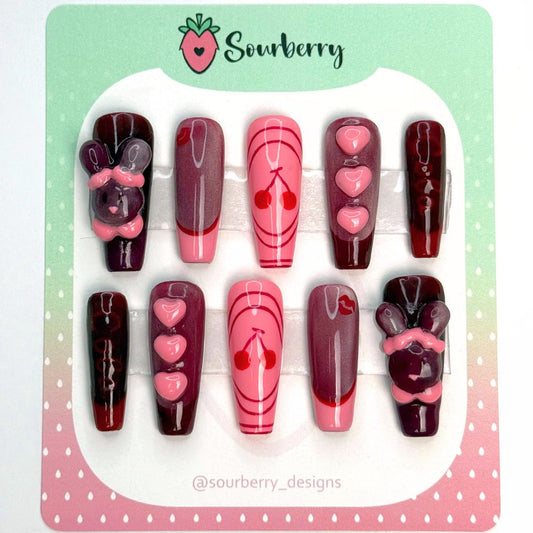 Cherry Cola nails. The thumb nail has a 3D sparkly red rabbit on it. The index nail is red and shimmery with a red kiss mark and a pink french tip. The middle nail is pink with a red cherry design. The ring nail is shimmery red with a dark red french tip and 3D pink hearts. The pinky nail is dark red with "cola" written on it and a red french tip.