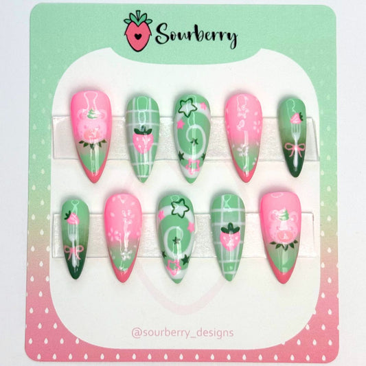 Strawberry Matcha Nails. The thumb nail is pink and green with a pink french tip and a light pink bear on it. The index nail is green with white stripes in the background and a pink strawberry in the middle. The middle nail is a green and white swirl with tiny pink and green stars. The ring nail is pink and green with light pink cherry blossom flowers on it. The pinky nail is light and dark green with a fork and a strawberry on it.