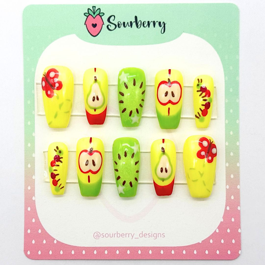 An image of Sourberry brand Fruit Basket press-ons mounted on a green and pink card. The nails are red, yellow, and green . The thumb nail has a butterfly, the index nail has a pear and a red french tip, the middle nail has a kiwi with stars, the ring nail has an apple and a green french tip, and the pinky nail has a multicolor caterpillar on it.