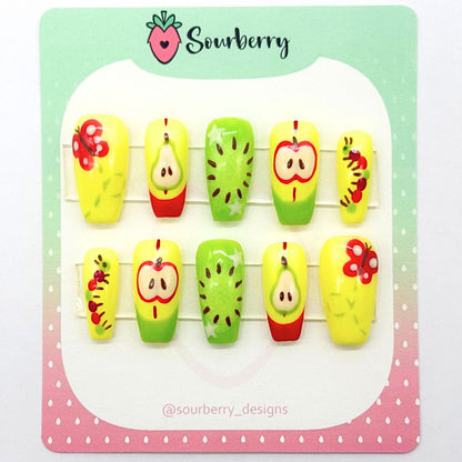 An image of Sourberry brand Fruit Basket press-ons mounted on a green and pink card. The nails are red, yellow, and green . The thumb nail has a butterfly, the index nail has a pear and a red french tip, the middle nail has a kiwi with stars, the ring nail has an apple and a green french tip, and the pinky nail has a multicolor caterpillar on it.