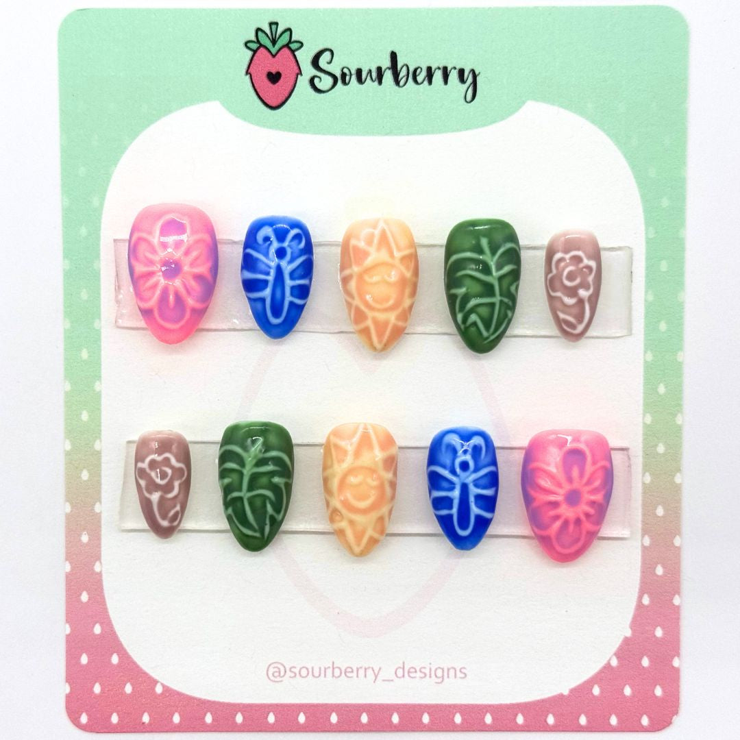 Sourberry brand Sunny Days press on set mounted on a pink and green card. The set has a ceramic tile effect. The thumb nail and pinky nails have flowers on them. The index nails have butterflies on them. The middle nails have suns on them. And the ring nails have a leaf on them.