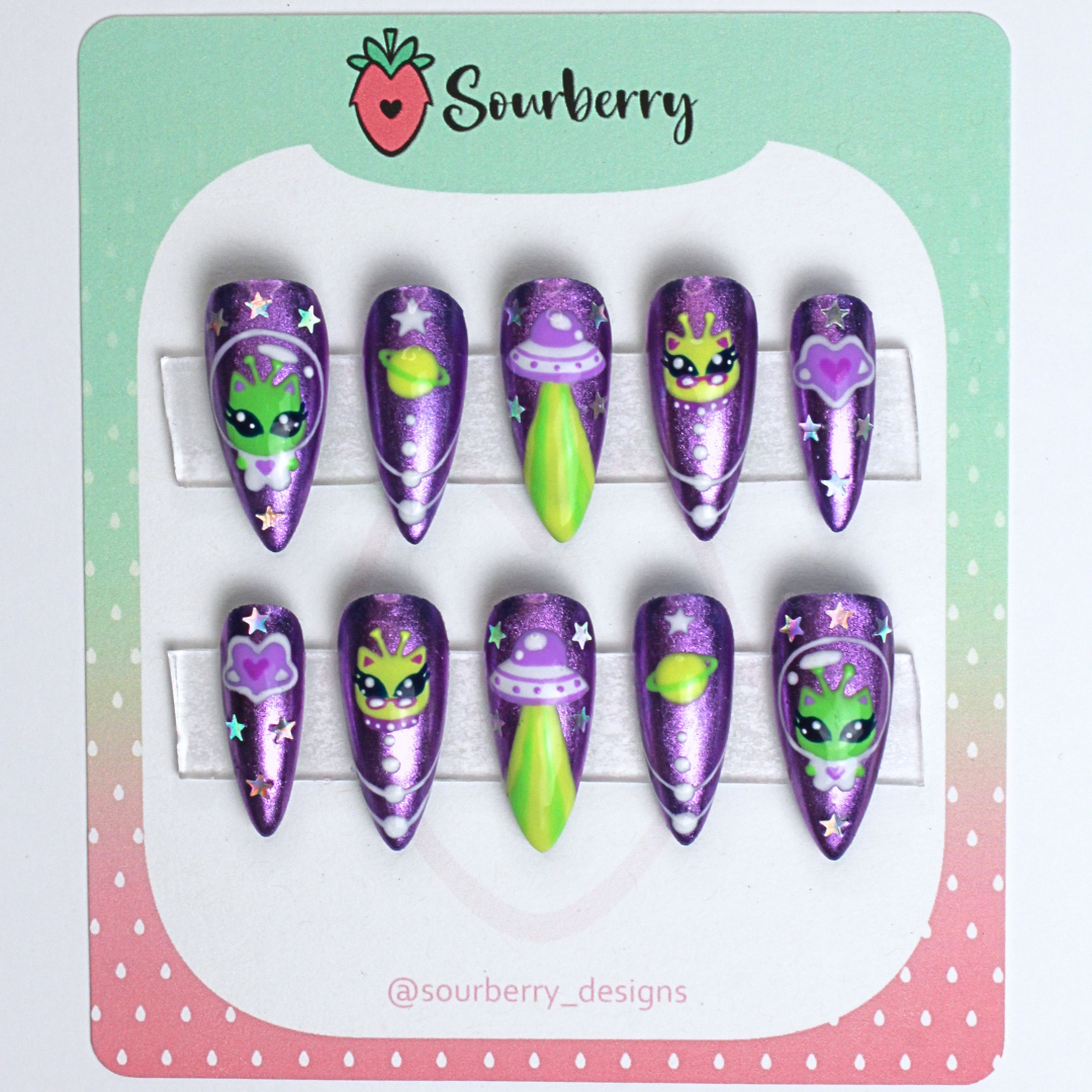 Galactic kitty nails mounted on a green and pink card. The nails are purple chrome with small paintings of alien kittens, planets, and stars on them.