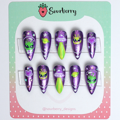 Galactic kitty nails mounted on a green and pink card. The nails are purple chrome with small paintings of alien kittens, planets, and stars on them.