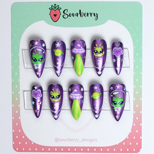 Galactic kitty nails mounted on a green and pink card. The nails are purple chrome with small paintings of alien kittens, planets, and stars on them.