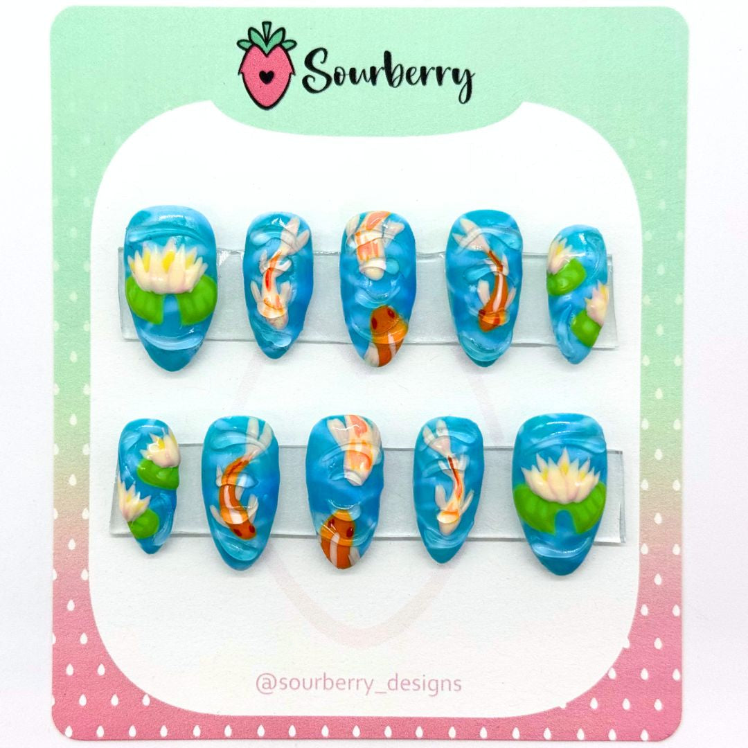 Sourberry Koi Pond nail set mounted on a green and pink card. The thumb nail and pinky nails have lotus flowers on them and all other nails have white and orange koi fish on them. They are topped with 3D water ripples.