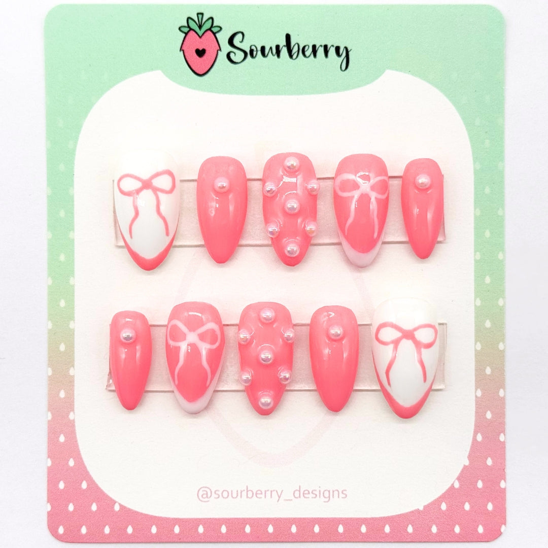 A picture of Sourberry brand "Pink Beau" press-on nails mounted on a green and pink card. The nails are white and pink. The thumb and ring finger nails have french tips and bows and the other nails have white pearl charms on them.