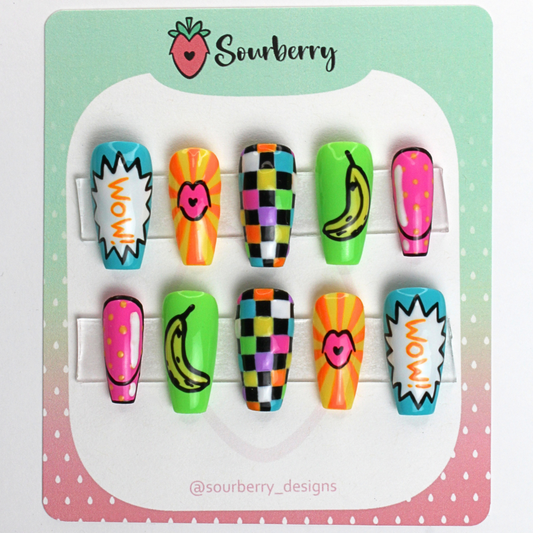 Pop Art nails. The thumb nail has a "wow" speech bubble. The index nail has cartoon lips. The middle nail has a rainbow and black checkerboard pattern. The ring nail has a cartoon banana. The pinky nail has a dotted pattern.