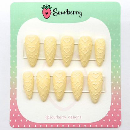 A picture of Sourberry brand "Powder Puff" press-on nails mounted on a green and pink card. The nails are light yellow with 3D heart and star patterns.