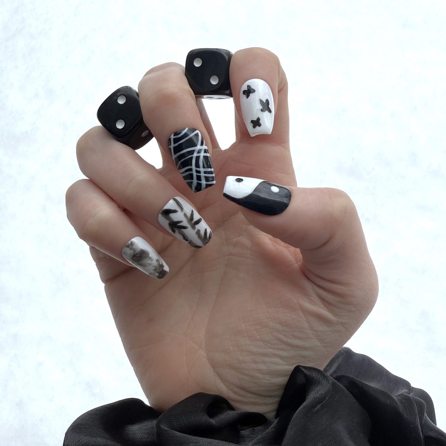 Yin Yang nails on a right hand. In between the first two fingers and the second two fingers are a pair of black dice. The background is white snow.