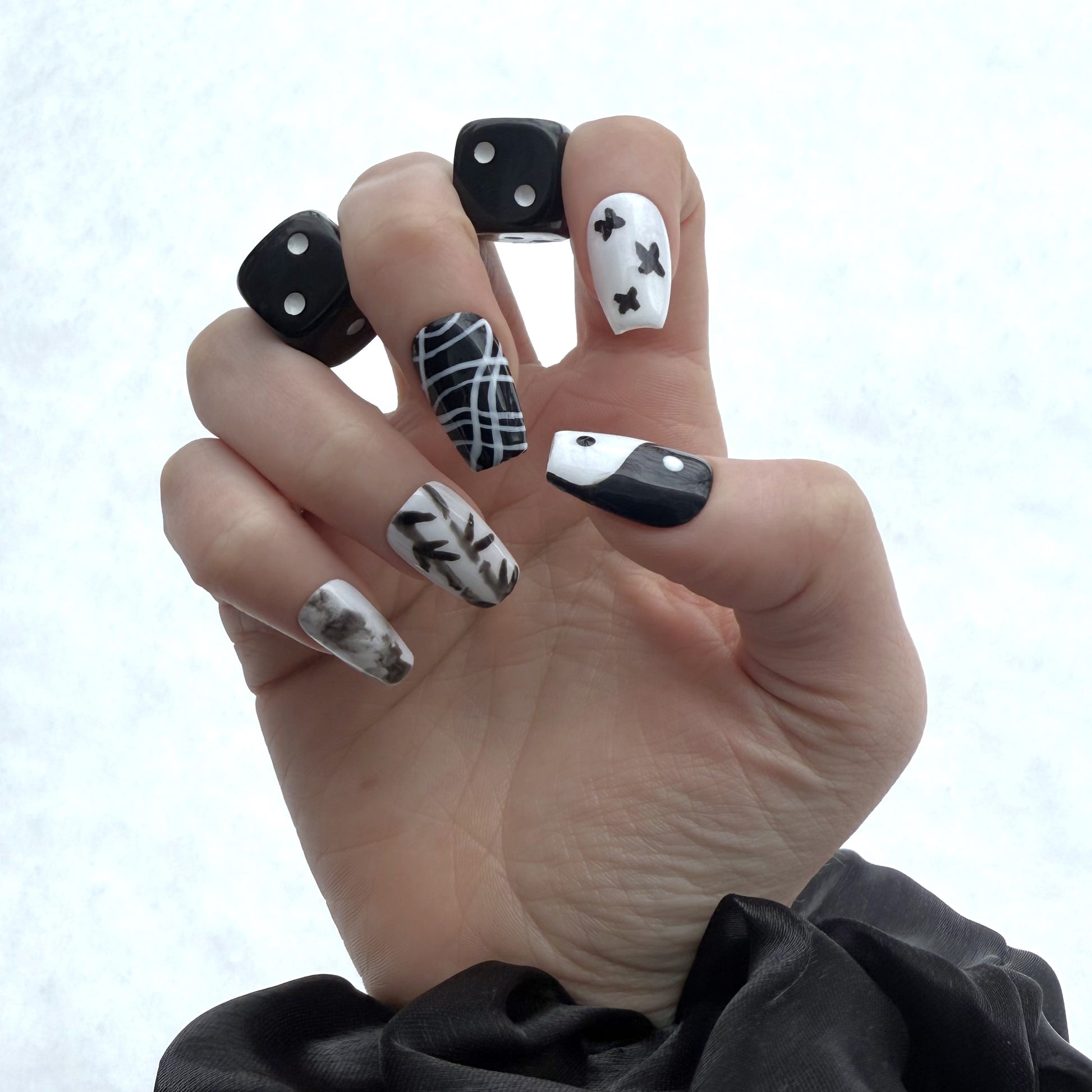 Yin Yang nails on a right hand. In between the first two fingers and the second two fingers are a pair of black dice. The background is white snow.