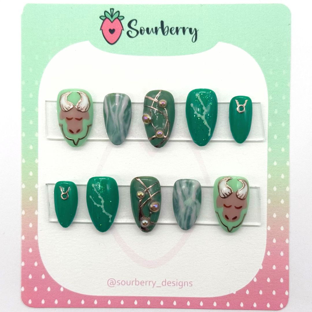 Sourberry brand Taurus nails mounted on a pink and green card/ The thumb nails have an image of a bull's head, the index nails are marbled with green and white, the middle nail has gold chrome details and brown pearls, the ring nail has glitter and the taurus constellation, the pinky nails have a golden taurus symbol.