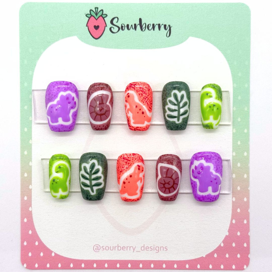 Sourberry brand tiny dinos nails. The thumb nail is purple with a triceratops, the index nail is brown with a fossil, the middle nail is orange with a t-rex, the ring nail is dark green with a fern, the pinky nail is light green with a brontosaurus.