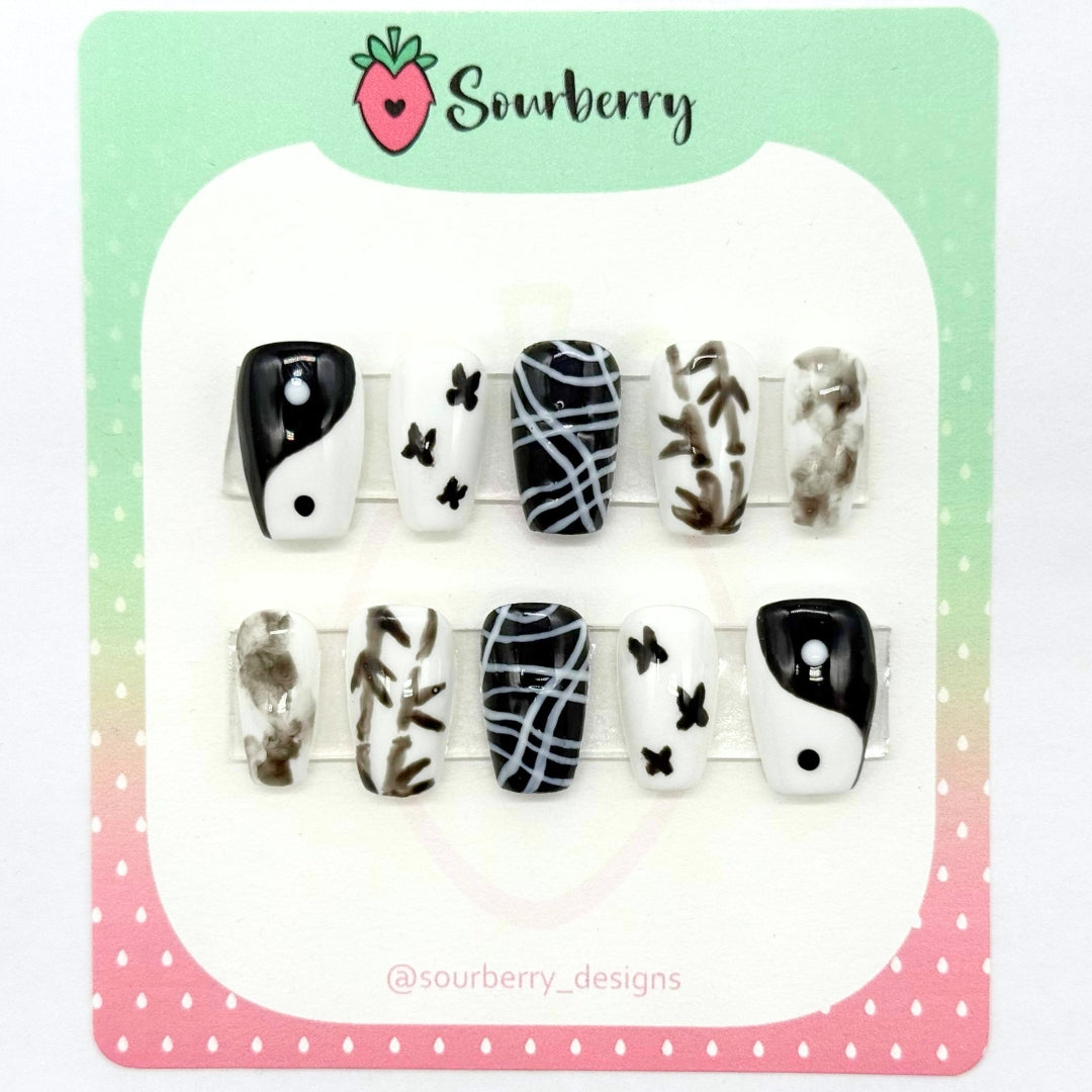 A picture of Sourberry brand "Yin Yang" press-on nails mounted on a green and pink card. The nails are white and black. The thumb nails have a yin-yang symbol. The index nails have three black butterflies on a white background. The middle nails have wavy white lines on a black background. The ring nails have black bamboo on them. And the pinky nails have a black ink pattern on them.